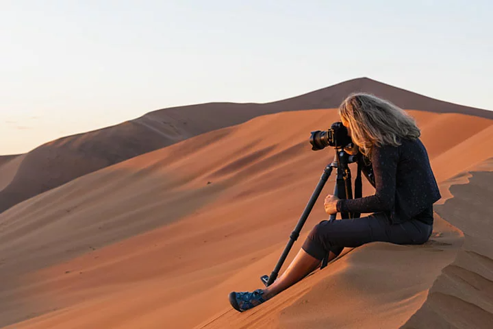 Una persona sentada sobre una duna en un desierto con una cámara sobre un trípode