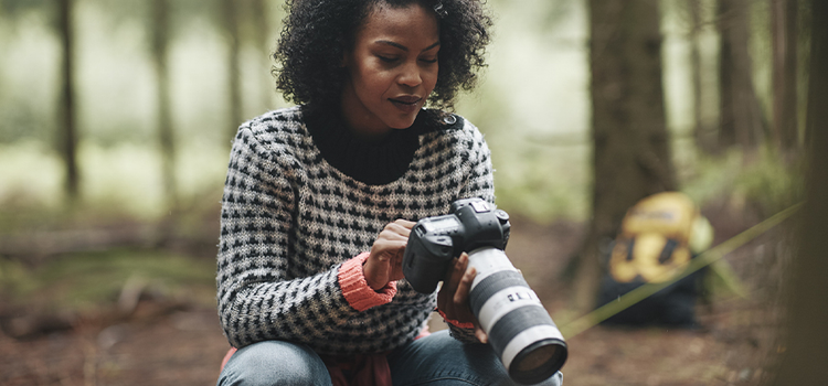 A person kneeling in a forest while looking at the exposure settings on their camera