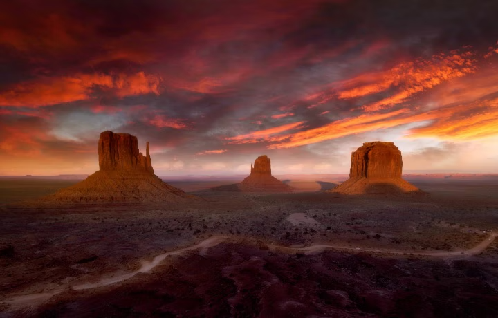 Three Monument Valley rock formations under dramatic red and orange sunset clouds in desert landscape