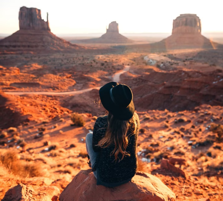 Woman with long hair and hat sitting on rock, viewed from behind, with three Monument Valley rock formations in the distance