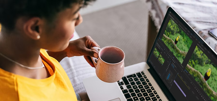 A person holding a coffee in one hand and transcoding a video with Adobe Premiere Pro on their laptop computer