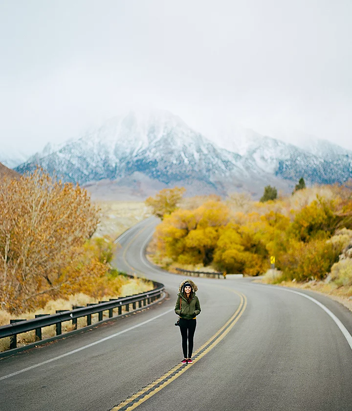 A person standing in the middle of a roadway leading to a snow-covered mountain range