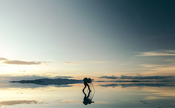 A photographer taking a photo with their camera on a tripod while standing in very shallow water