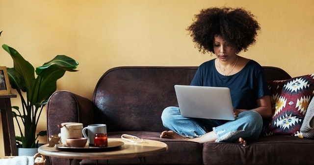 A photo of a person sitting cross-legged on a couch and using a laptop computer on their lap.