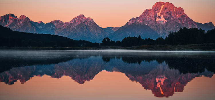 A body of water reflecting an image of a forest and a snow-covered mountain range behind it