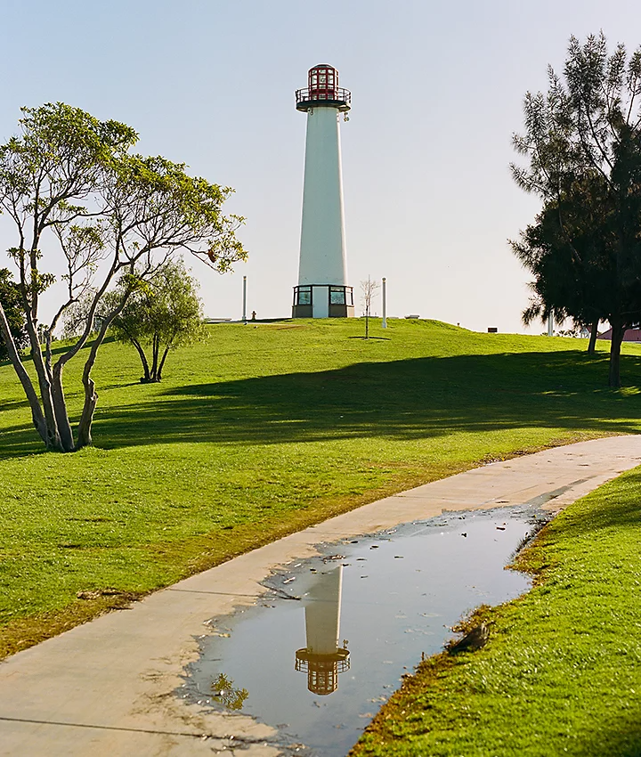 A lighthouse on a small grassy hillside and its reflection in a water puddle in front of it