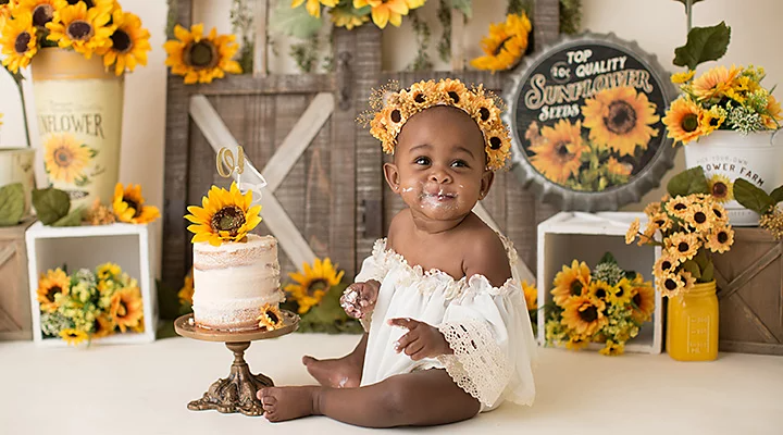 A baby sitting next to a cake surround by various props that are sunflower-themed