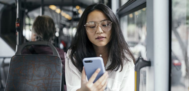 A person wearing glasses sits on a bus looking at a mobile device