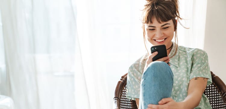 A person smiling and sitting in a wicker chair while looking at a mobile device