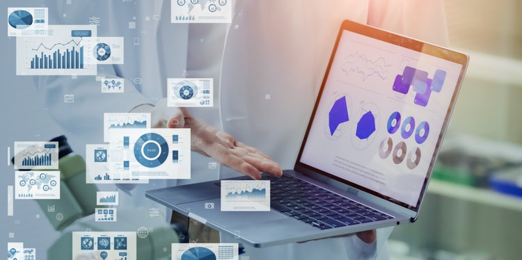 A person in white jacket holding and pointing at laptop screen with dynamic data and graphs.