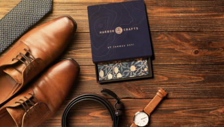 Close up of brown and black shoes, tie, belt, watch, and box layered over realistic wood floor of browns and black wood grain.