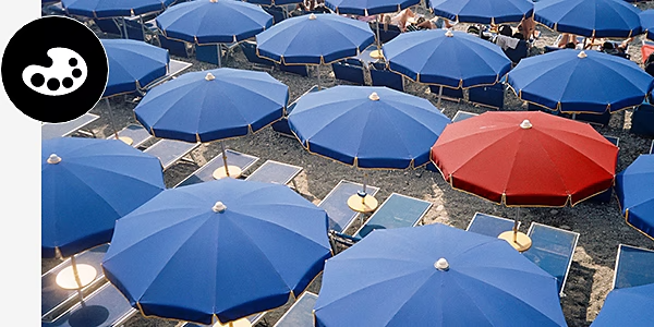 An image of sun umbrellas on the beach. Most of the umbrellas are blue but one umbrella is red.