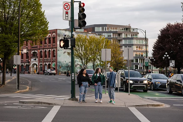 A photo of three people standing at an intersection, waiting to cross the street.