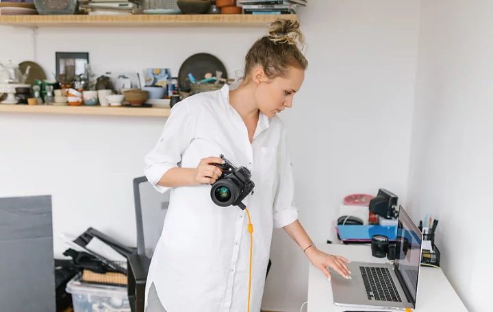 A photo of a photographer standing in their office with their camera in one hand, using a laptop on a desk with their other hand.