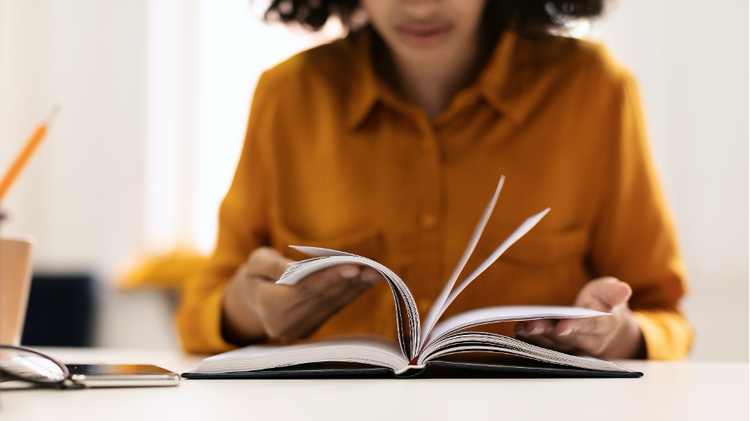 Photo of a young woman sitting at a table with a book, pen, glasses and smartphone.