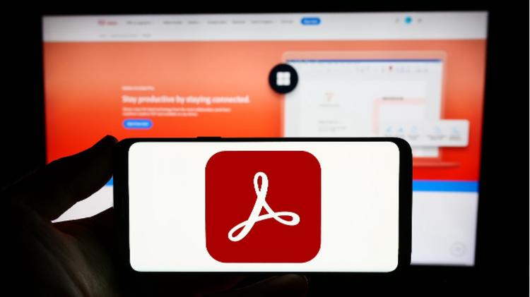 Photo of a person holding a smartphone with the Adobe Acrobat logo displayed on the screen. In the background is a computer screen with the page for Adobe Acrobat Online.