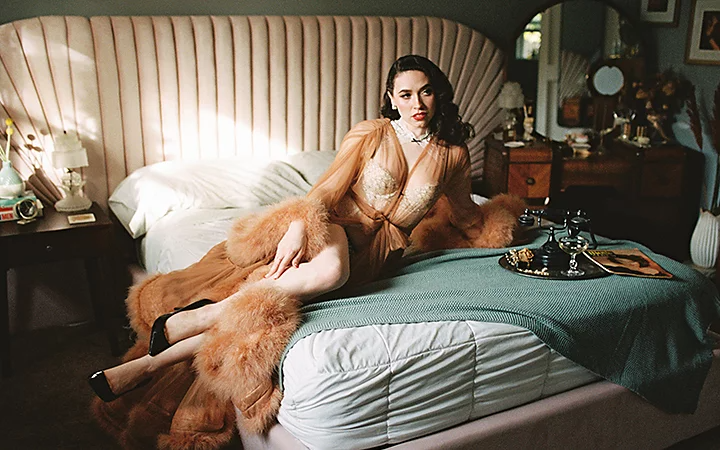 A person posing in vintage clothes on a bed next to a tray with a vintage rotary phone on it