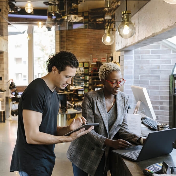 Woman in business attire and a man in a black shirt reviewing business files on laptop and tablet