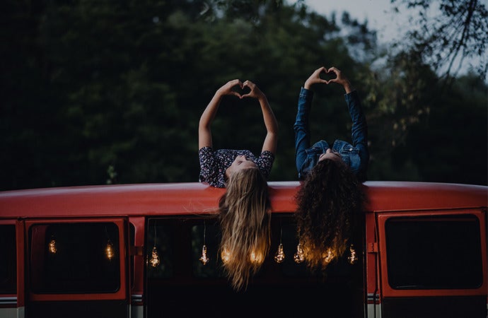 Two girls laying on top of a camping van making hearts with their fingers during blue hour.