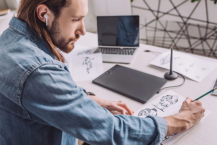 Man sat at desk with laptop drawing different animated characters.