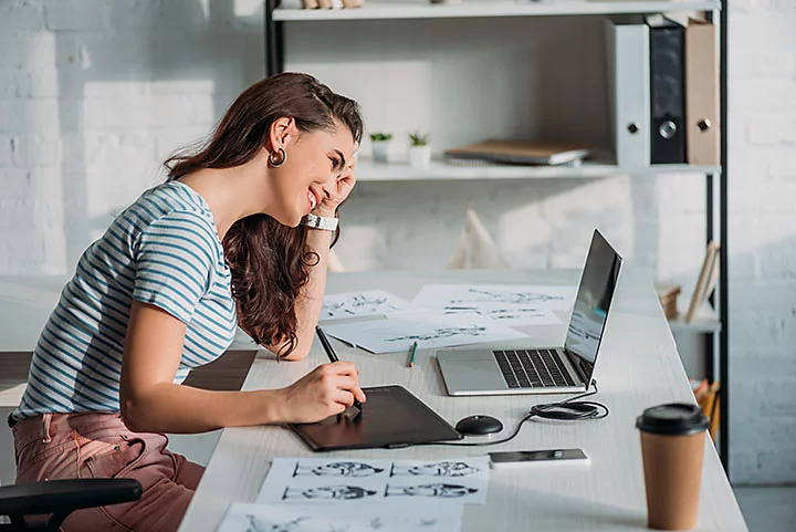 Woman at desk looking at laptop drawing on design pad.