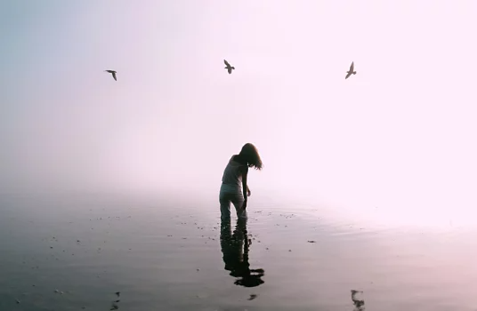 A person standing in shallow water as three birds fly above them