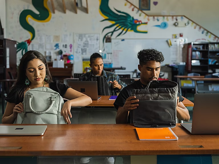 three people sit in a classroom with bags, laptops and notebooks