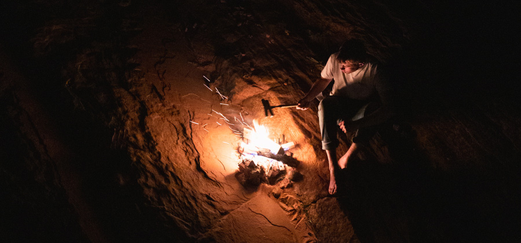 A high-contrast photo of a person cooking hot dogs over a campfire at night